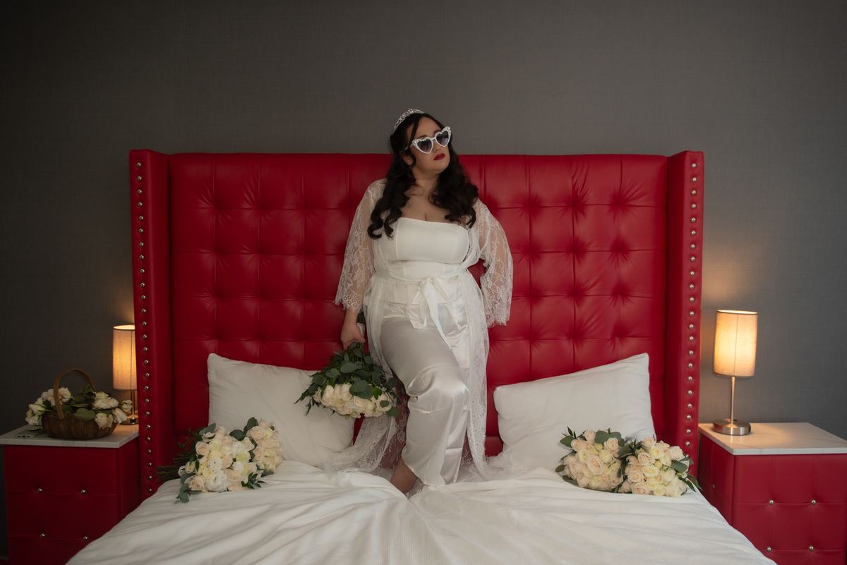 A bride in a white satin and lace robe, heart-shaped sunglasses, and a tiara poses on a red bed with white roses.