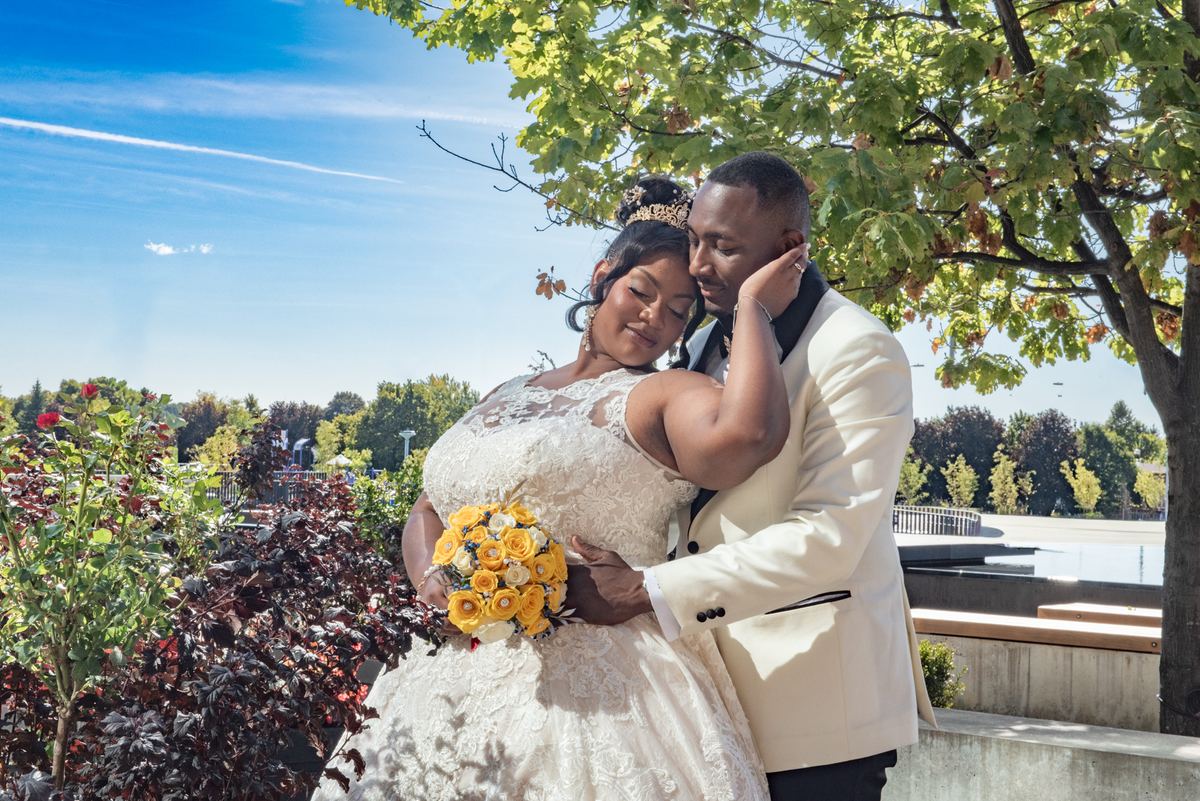 A bride in a lace gown and yellow roses rests her head on her groom's shoulder under leafy trees, both smiling.
