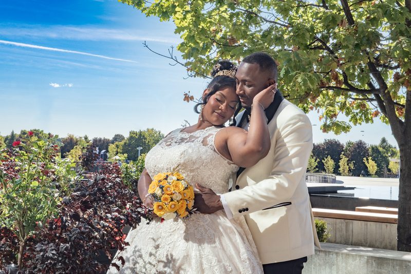 A bride in a lace gown and yellow roses rests her head on her groom's shoulder under leafy trees, both smiling.