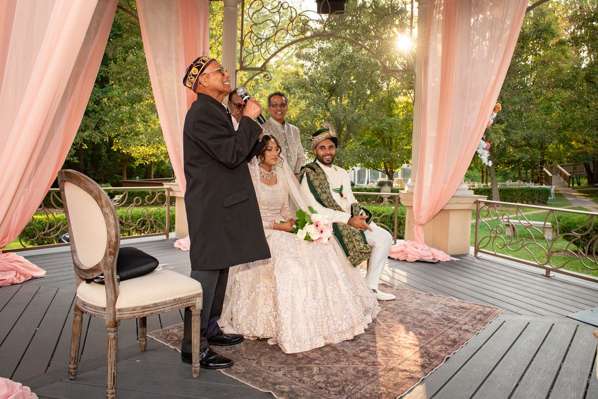 A bride and groom sit under a decorated gazebo during an outdoor ceremony, with guests smiling in the background.