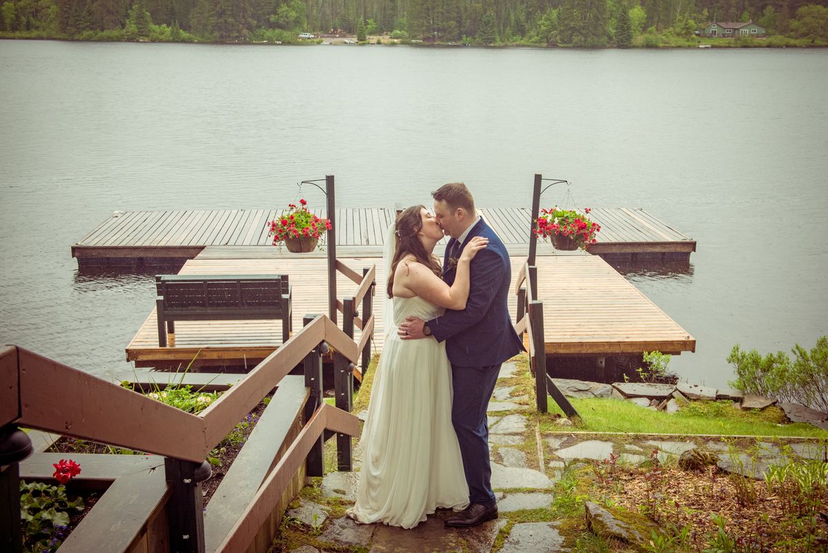 A bride and groom kiss beside a wooden dock over a calm lake, surrounded by hanging flower baskets and forested shore.