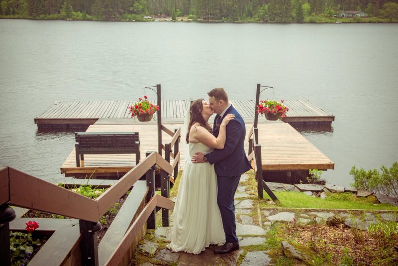 A bride and groom kiss beside a wooden dock over a calm lake, surrounded by hanging flower baskets and forested shore.