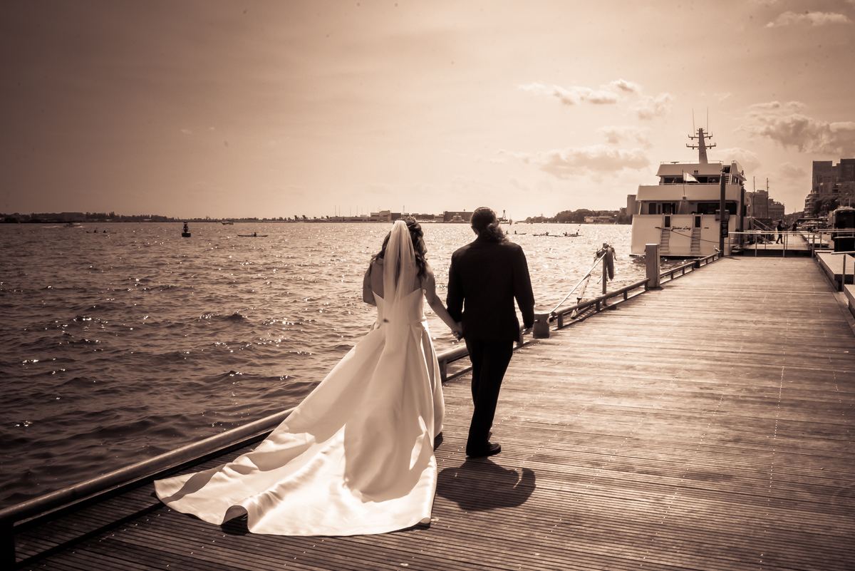 A bride and groom walk hand-in-hand along a wooden pier, bathed in warm golden light reflecting on the water.
