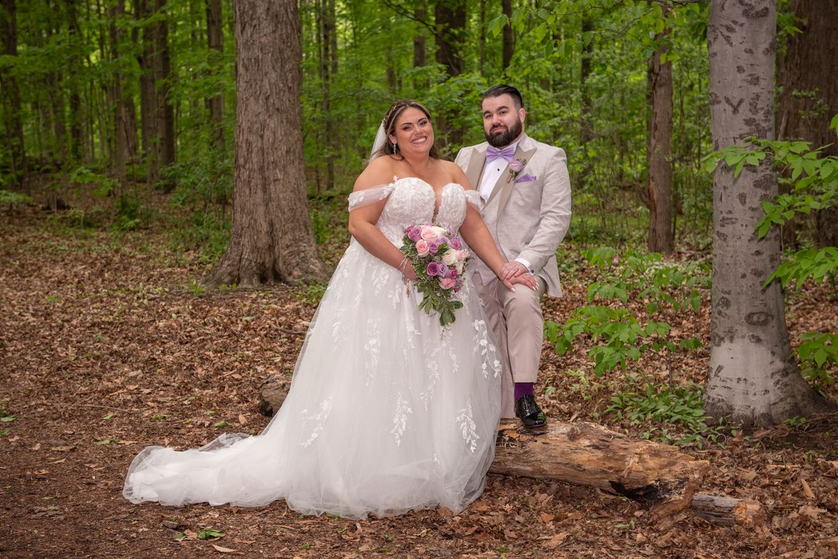 A bride in a white gown and groom in a gray suit with a purple bow tie pose happily in a sunlit forest clearing.