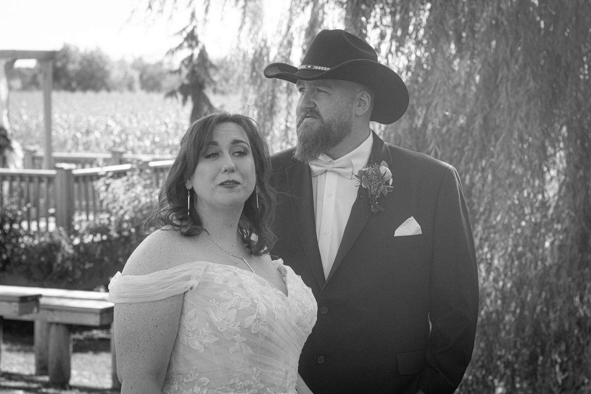 Bride in an off-the-shoulder lace gown and groom in cowboy hat stand outdoors under dappled sunlight.