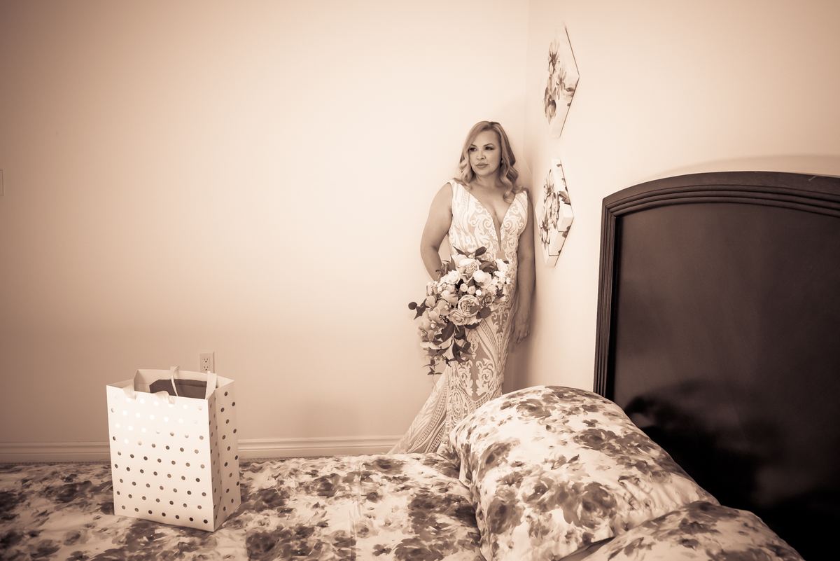 A bride stands in a softly lit bedroom, leaning against the wall beside a floral-patterned bed with her bouquet.