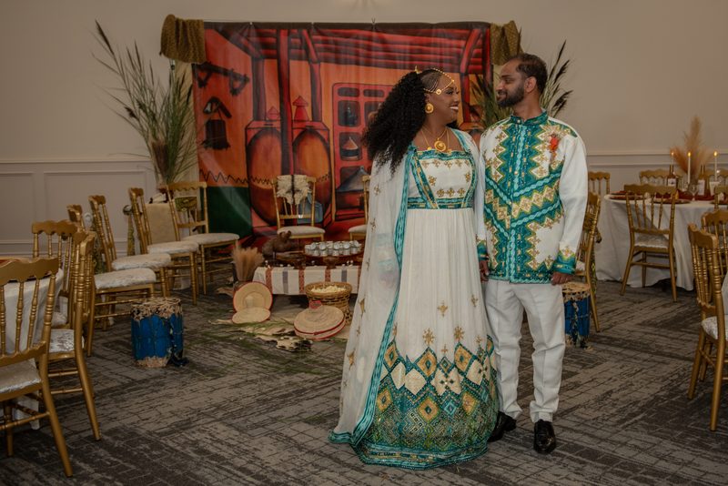 A bride and groom in elaborate traditional African attire smile at each other in a hall dressed with drums and gold chairs.