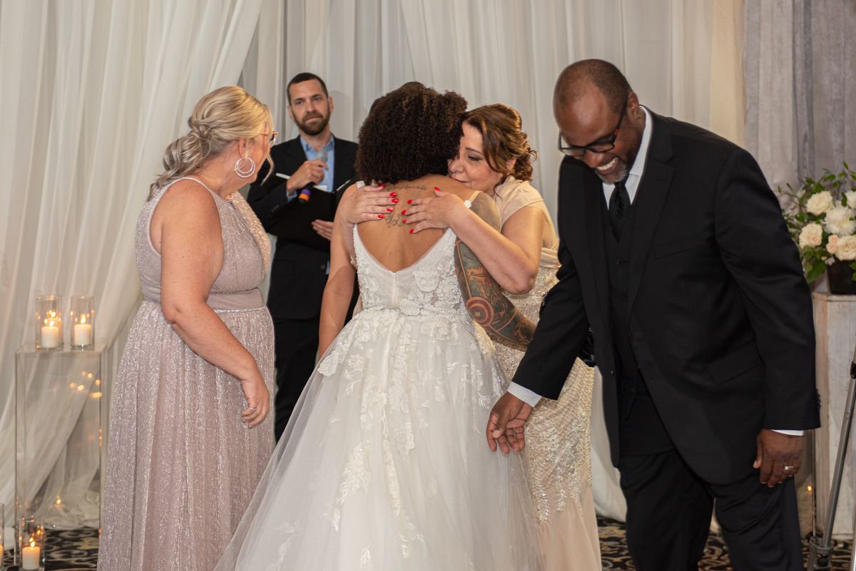 A bride is embraced by a woman with tattoos while holding a man's hand, near two other suited men and a sparkly-gowned woman.