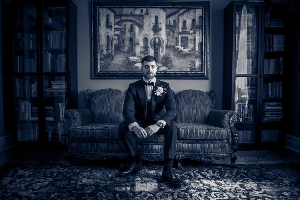 A groom in a tuxedo sits solemnly on an ornate sofa, bathed in cool blue light, surrounded by books.