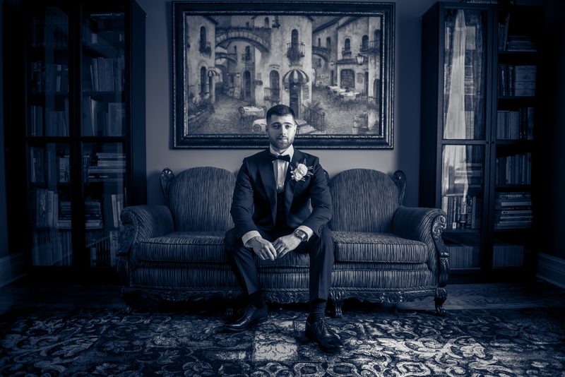 A groom in a tuxedo sits solemnly on an ornate sofa, bathed in cool blue light, surrounded by books.