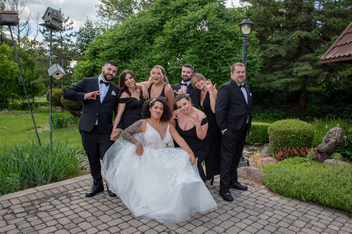A bride and groom pose playfully with their bridal party on a stone pathway surrounded by lush greenery.