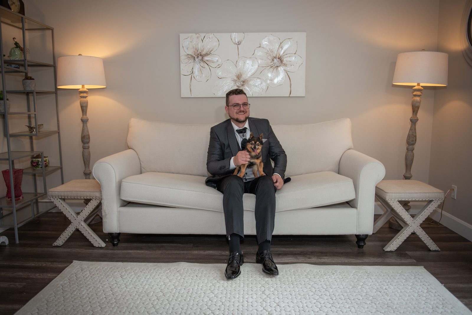 A man in a suit sits on a white sofa with a small dog, surrounded by neutral tones and decorative elements.