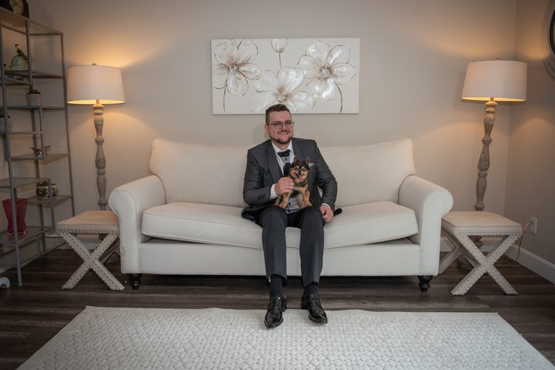 A man in a suit sits on a white sofa with a small dog, surrounded by neutral tones and decorative elements.