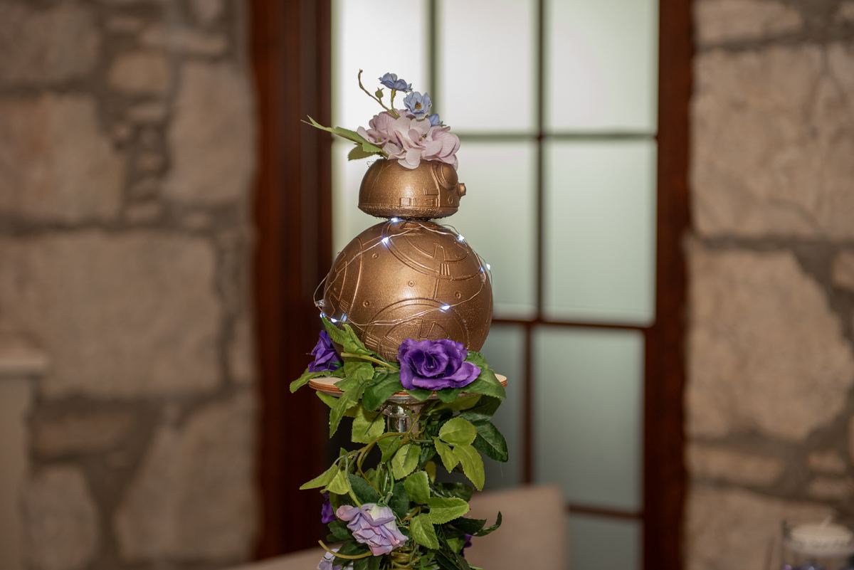 A decorated BB-8 droid with flowers and lights stands as a centerpiece at an event near a stone wall and glass door.