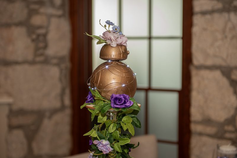 A decorated BB-8 droid with flowers and lights stands as a centerpiece at an event near a stone wall and glass door.
