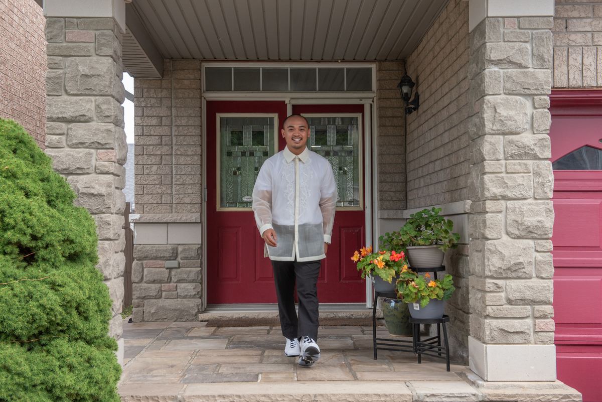 A man in a Barong Tagalog and dark pants walks toward the camera on a stone porch with red door and columns.