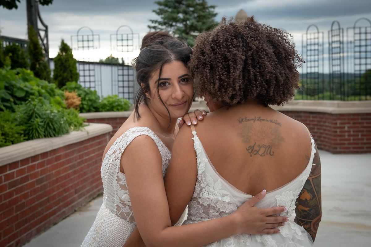 Two brides embrace tenderly outdoors, one smiling at the camera while the other's tattoo is visible on her back.