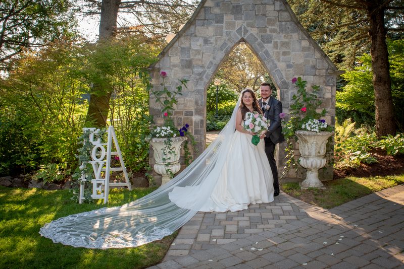 A bride and groom stand beneath a stone Gothic archway in a sun-dappled garden, the bride's veil trailing on the grass.