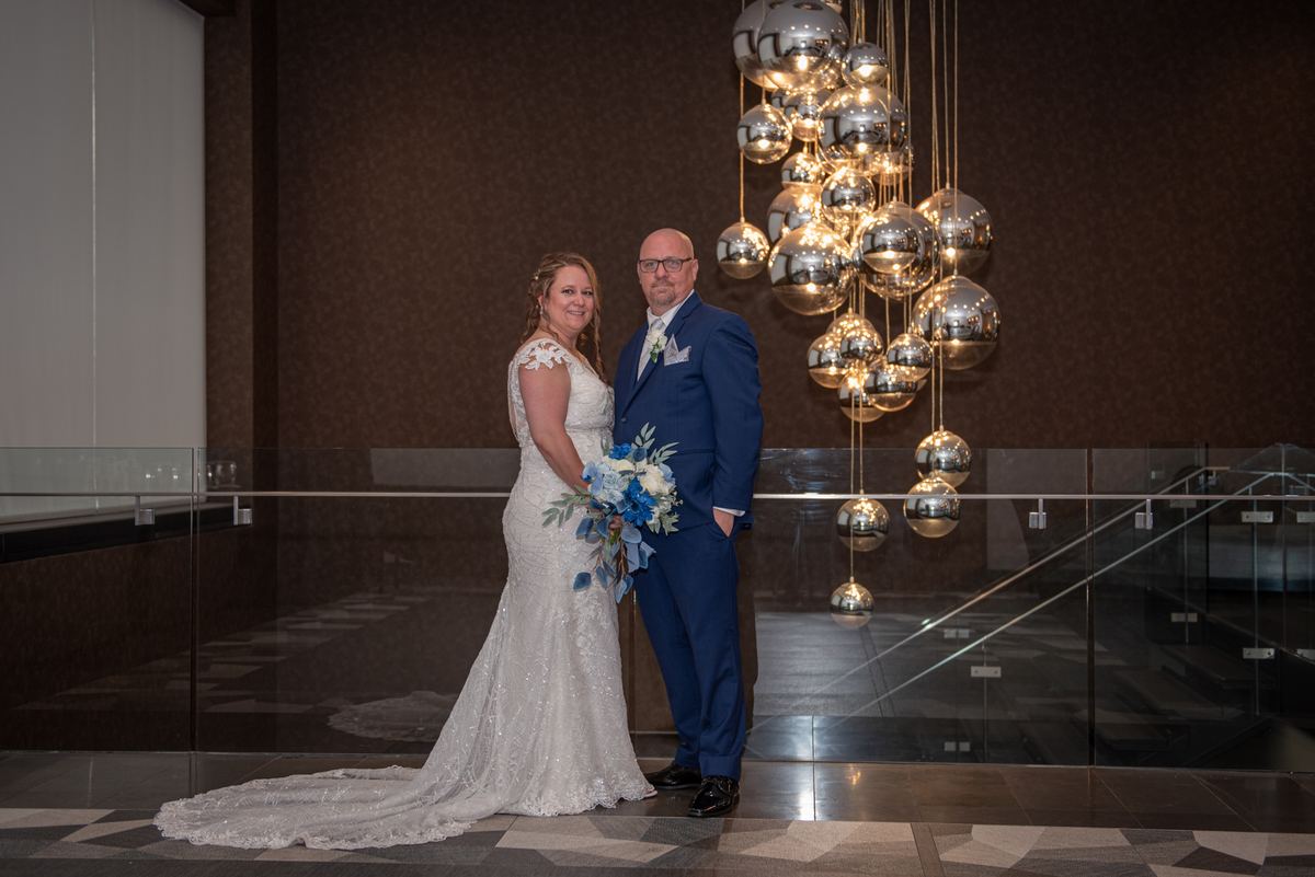 A bride and groom smile in a modern atrium, surrounded by a chandelier of mirrored orbs reflecting warm light.