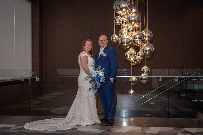 A bride and groom smile in a modern atrium, surrounded by a chandelier of mirrored orbs reflecting warm light.
