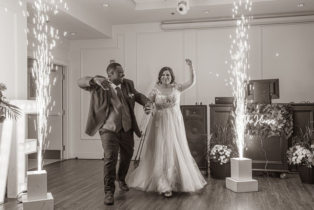 A joyful bride and groom dance amidst sparkling fireworks from pedestals in a decorated indoor space with event equipment.