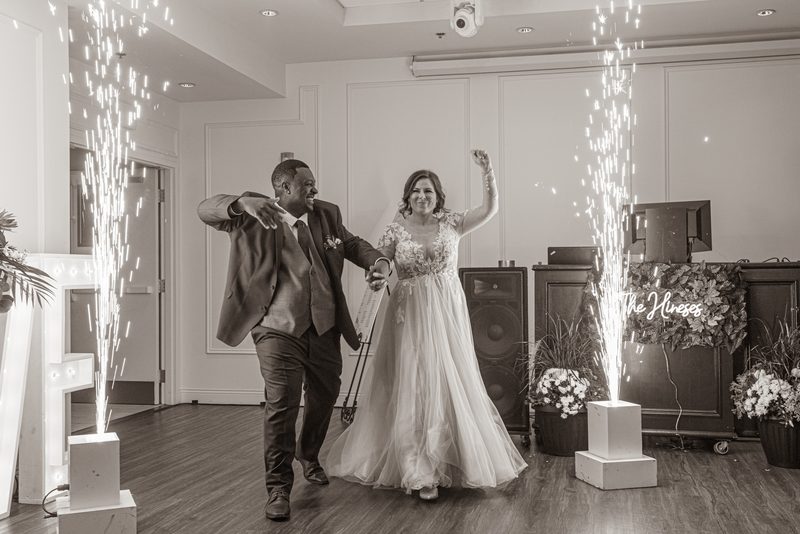 A joyful bride and groom dance amidst sparkling fireworks from pedestals in a decorated indoor space with event equipment.