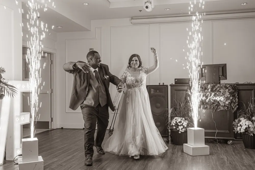 A joyful bride and groom dance amidst sparkling fireworks from pedestals in a decorated indoor space with event equipment.