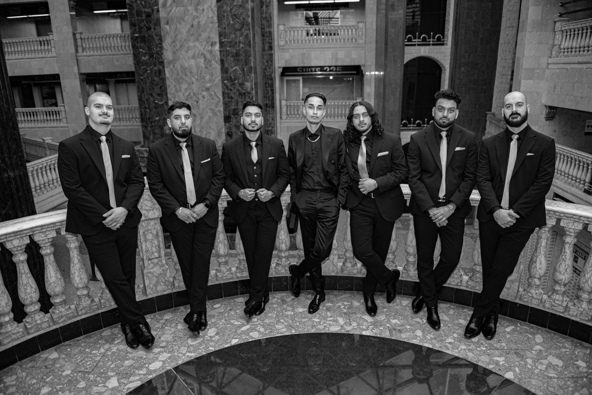 Seven groomsmen in black suits stand confidently in an ornate atrium with marble floors and railings.