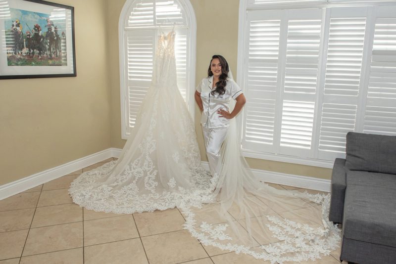 A bride in pajamas smiles next to her wedding gown on a hanger near white shutters, lit by soft daylight.