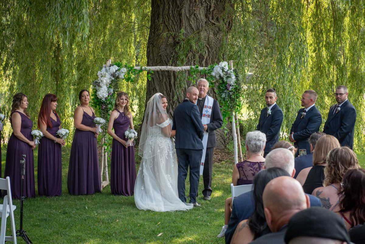 A bride and groom stand under a floral arch in a natural setting, surrounded by guests and their bridal party.