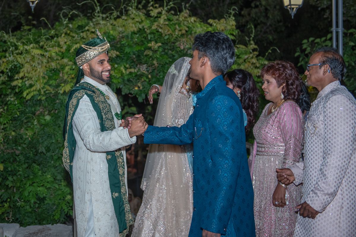 A groom in a white and green sherwani shakes hands with an elder as the bride in a white lehenga smiles beside them.