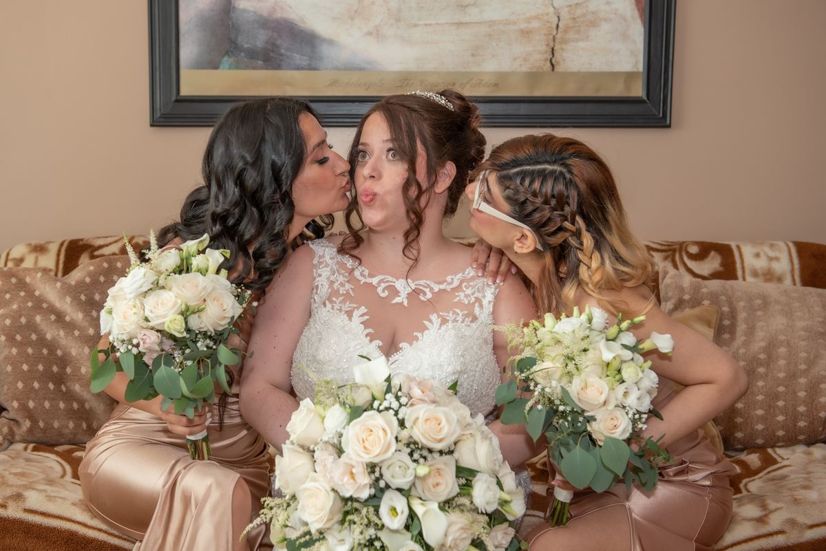 A bride and two bridesmaids share affectionate kisses while sitting on a couch with floral bouquets.