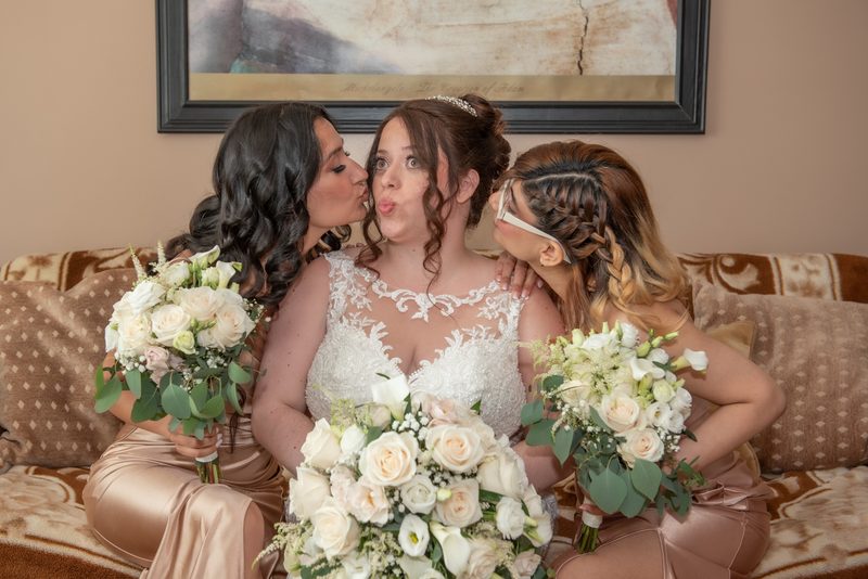 A bride and two bridesmaids share affectionate kisses while sitting on a couch with floral bouquets.