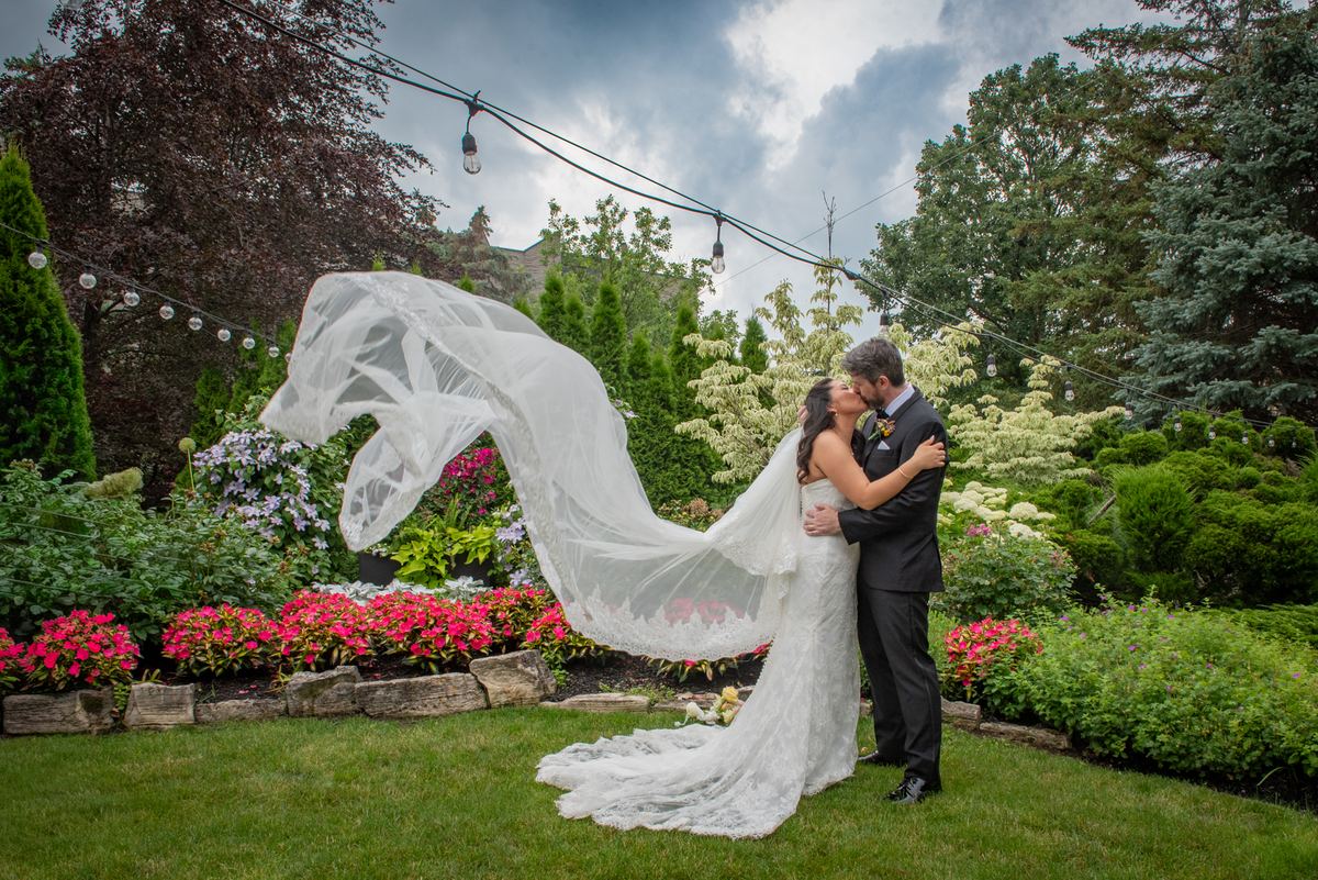 A bride and groom kiss in a garden with wind-blown veil and hanging lights aglow against the cloudy sky.