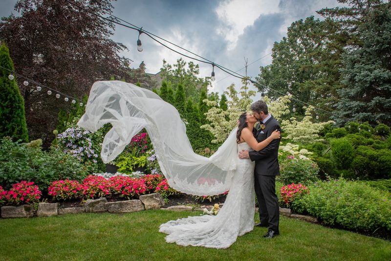 A bride and groom kiss in a garden with wind-blown veil and hanging lights aglow against the cloudy sky.
