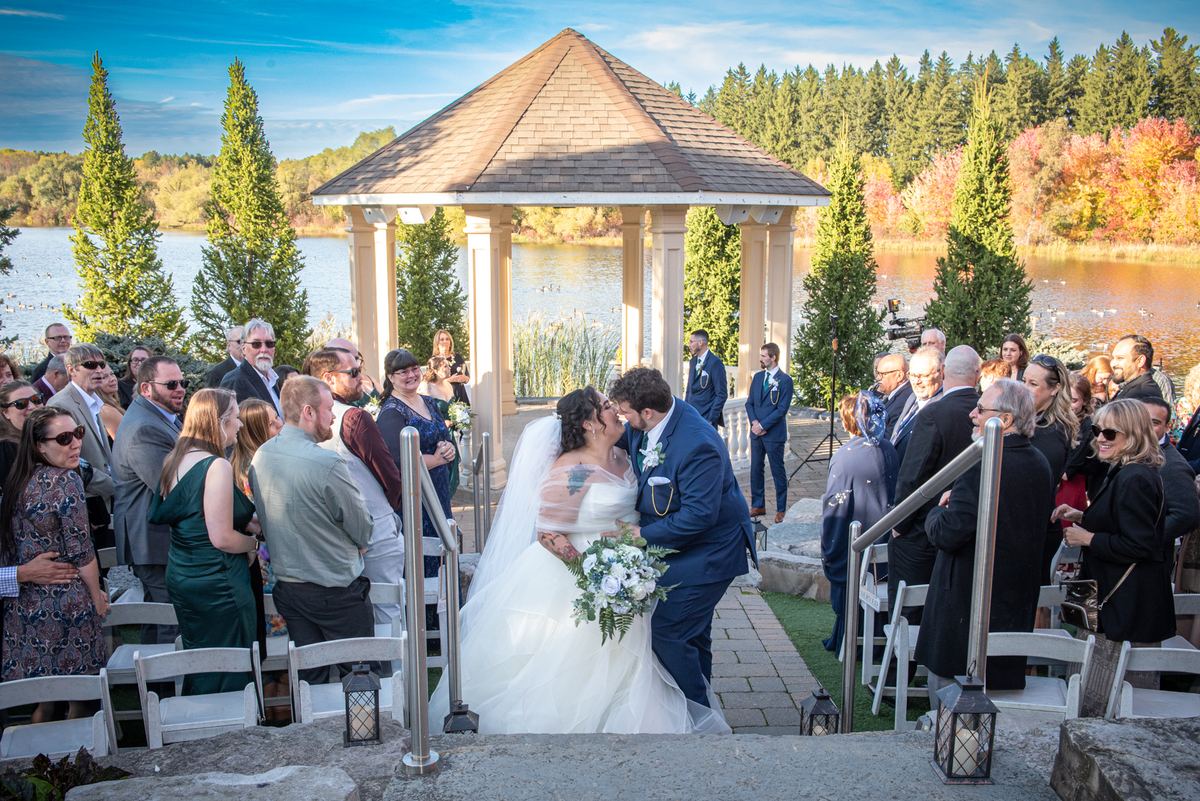 A bride and groom kiss under a wooden gazebo during an outdoor wedding, surrounded by guests and autumn scenery.