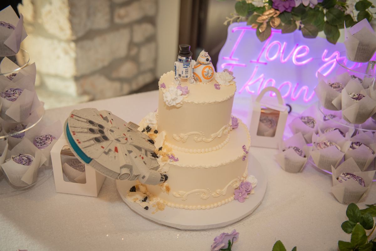 A wedding cake with Star Wars toppers and a Millennium Falcon model under a purple neon sign, surrounded by cupcakes.