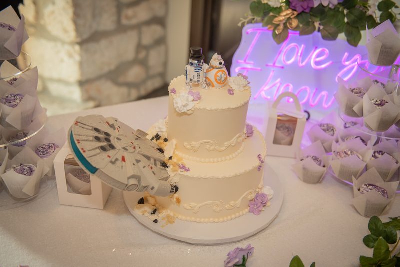 A wedding cake with Star Wars toppers and a Millennium Falcon model under a purple neon sign, surrounded by cupcakes.