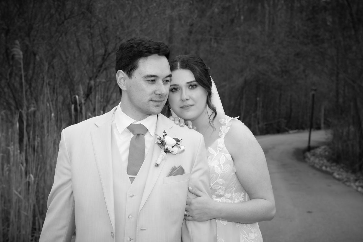 A bride in a lace gown and veil stands closely with her groom beside reeds, hand resting on his chest.