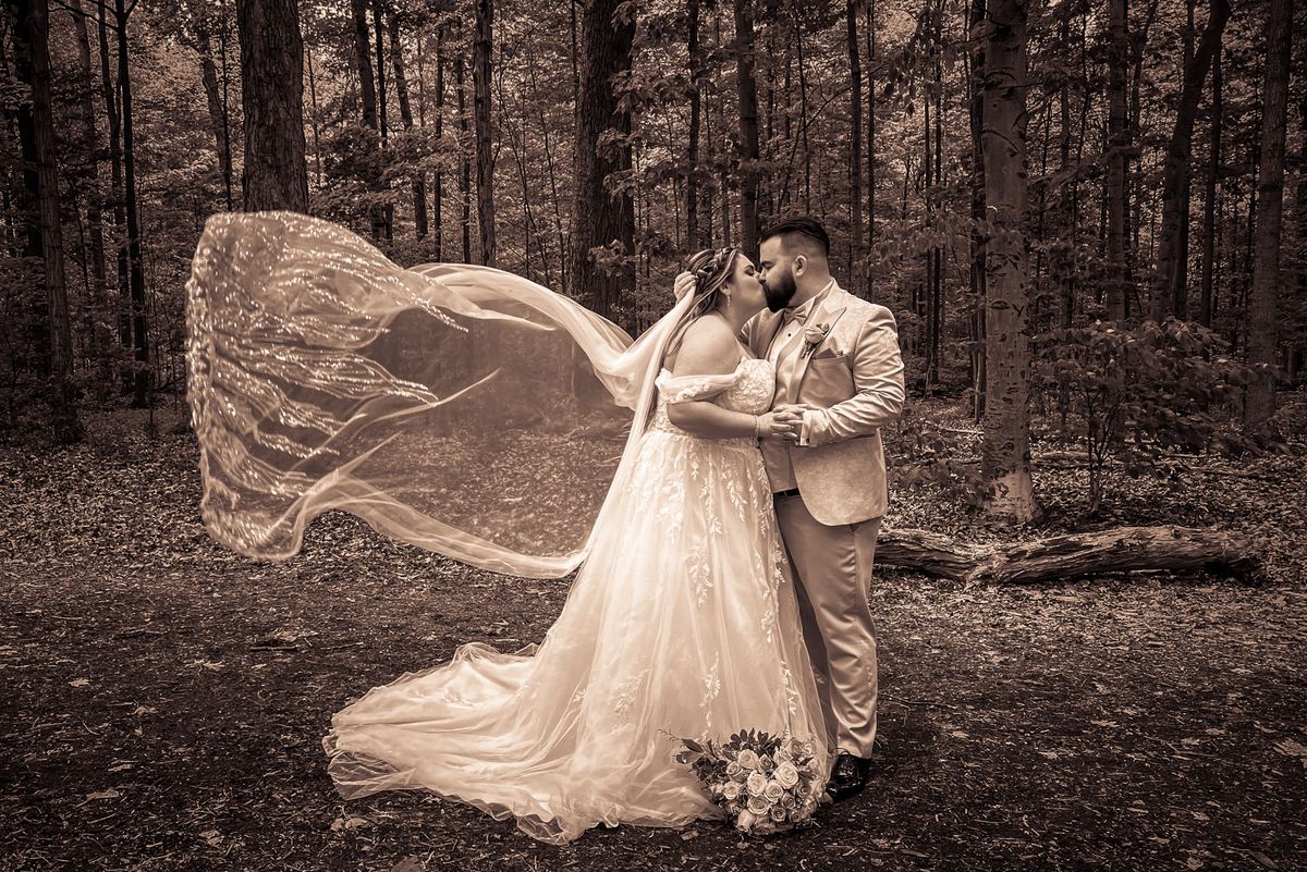 A bride and groom kiss tenderly in a forest, their long veil billowing in the wind as soft light filters through the trees.