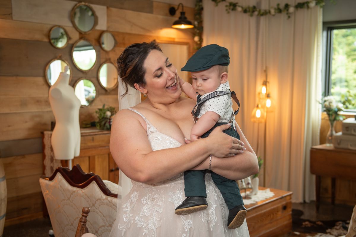 A bride in her wedding gown holds her child in a rustic indoor setting with wooden walls and windows.