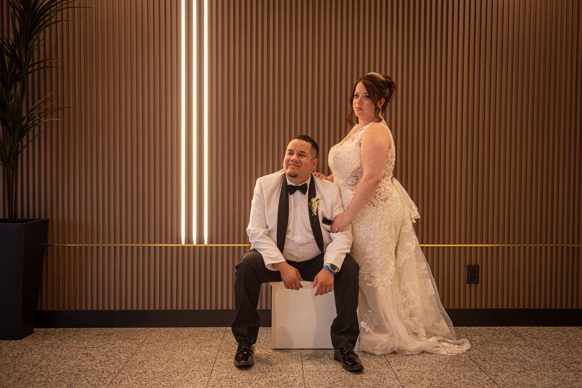 A bride and groom pose indoors, her hand on his shoulder near a wooden wall with vertical light strips.