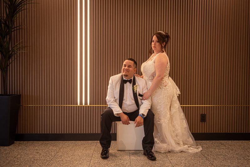 A bride and groom pose indoors, her hand on his shoulder near a wooden wall with vertical light strips.