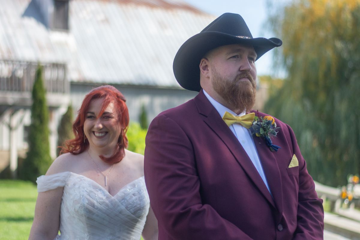 A smiling red-haired bride walks beside her groom in a maroon suit and black cowboy hat towards an outdoor ceremony.