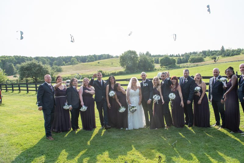 A joyful wedding party poses on a grassy field with the bride and groom at center, surrounded by flying kites.