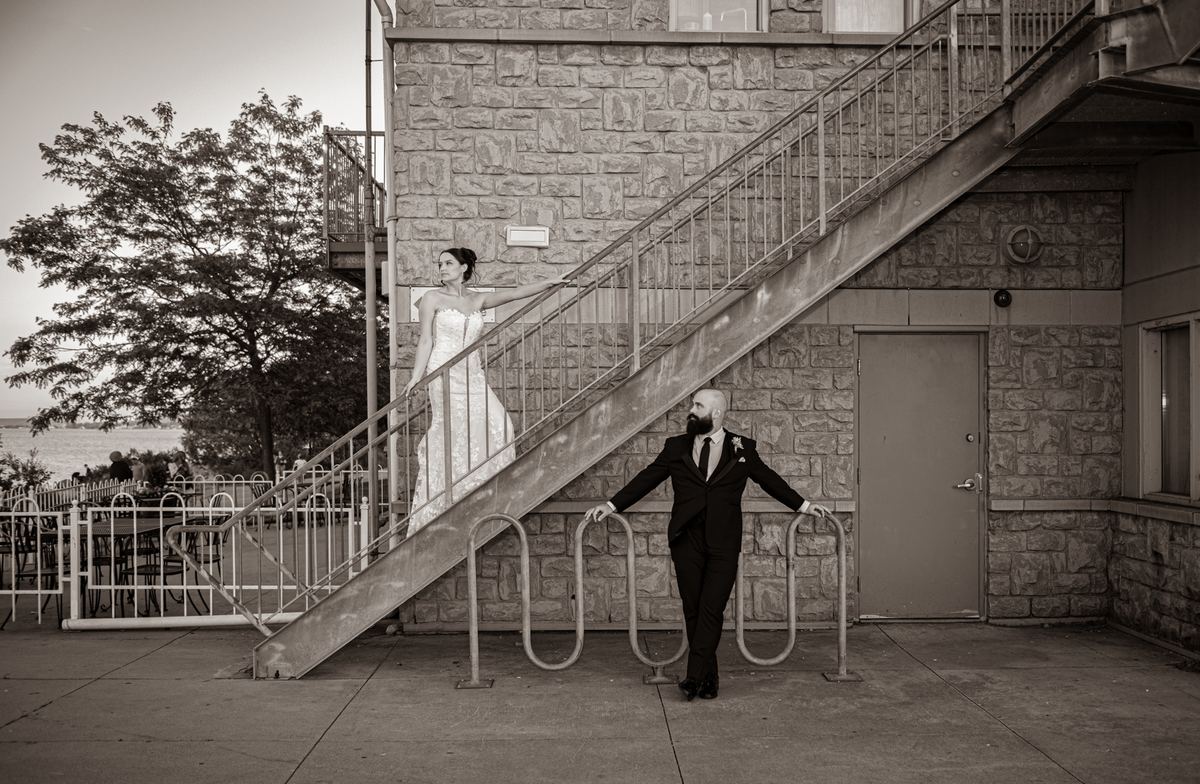 A bride and groom pose dramatically on a stone staircase near a bike rack, with a tree and waterfront in the background.
