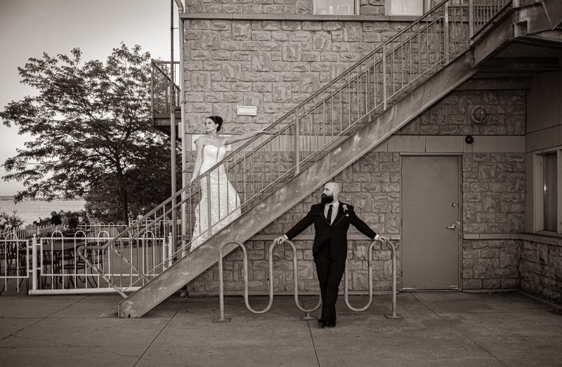 A bride and groom pose dramatically on a stone staircase near a bike rack, with a tree and waterfront in the background.
