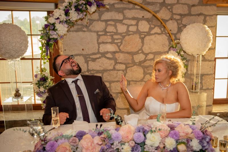 A laughing groom and amused bride sit at their reception table under warm light with floral decorations.