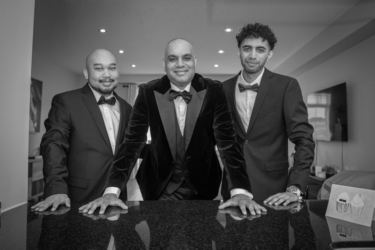 Three men in tuxedos smile and lean on a glossy countertop, illuminated by soft indoor lighting with reflections visible.