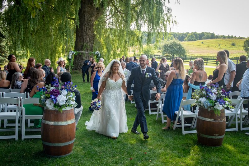 A smiling bride and groom walk hand-in-hand down a grassy aisle lined with wooden barrels and flowers, surrounded by guests.